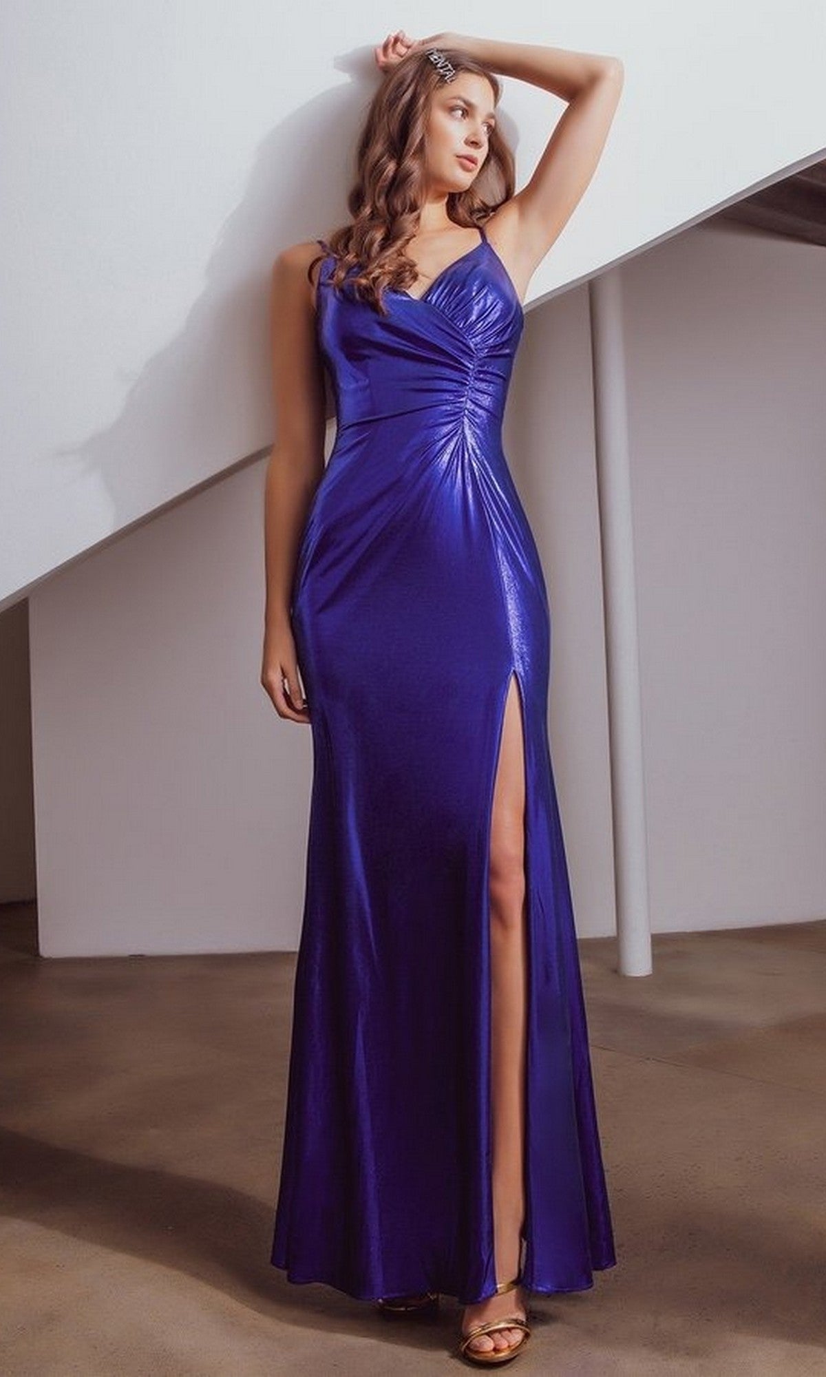 A woman stands indoors in the Eureka 9030 long prom dress in royal, a form-fitting lamé foil jersey gown with a v-neckline and thigh-high slit, paired with gold heels, her hand resting on her head.